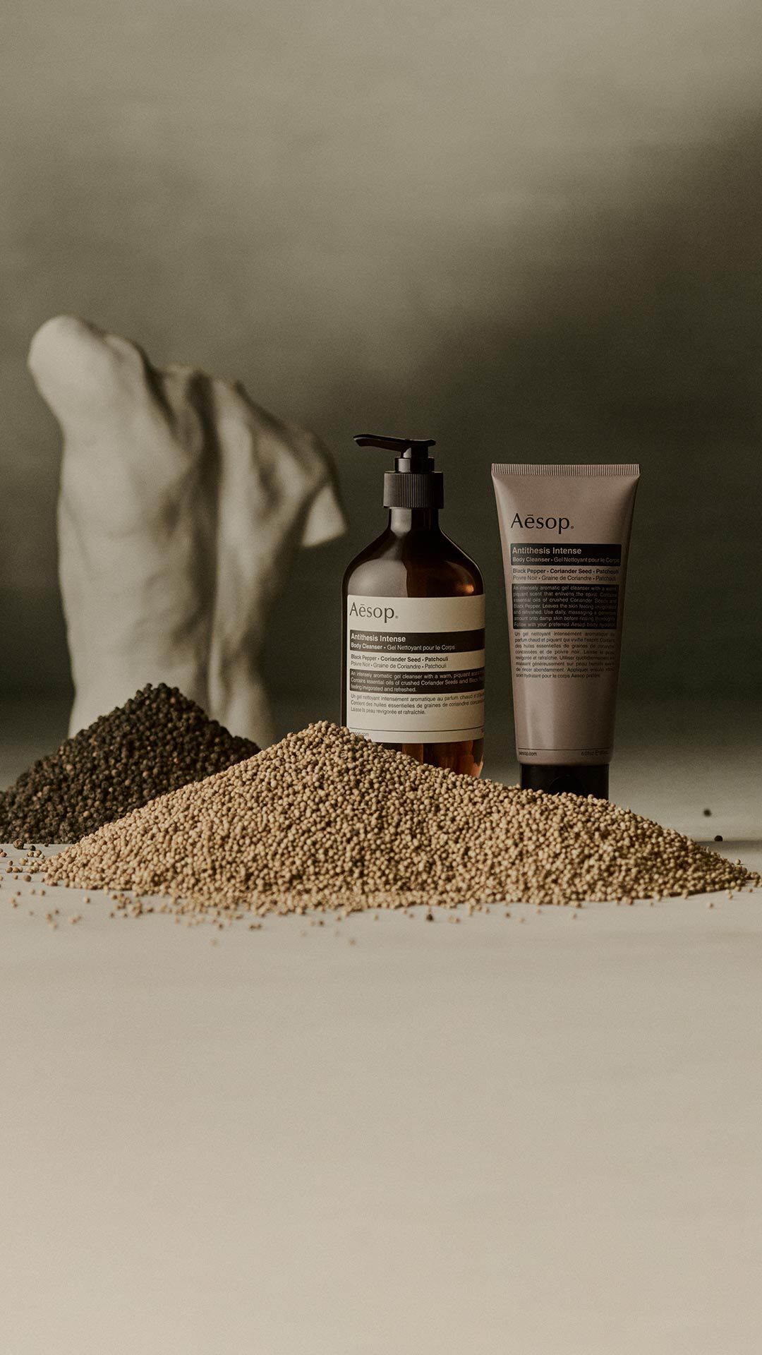 Aesop Antithesis Intense Body Cleanser pump bottle and tube beside piles of black peppercorns and coriander seeds.