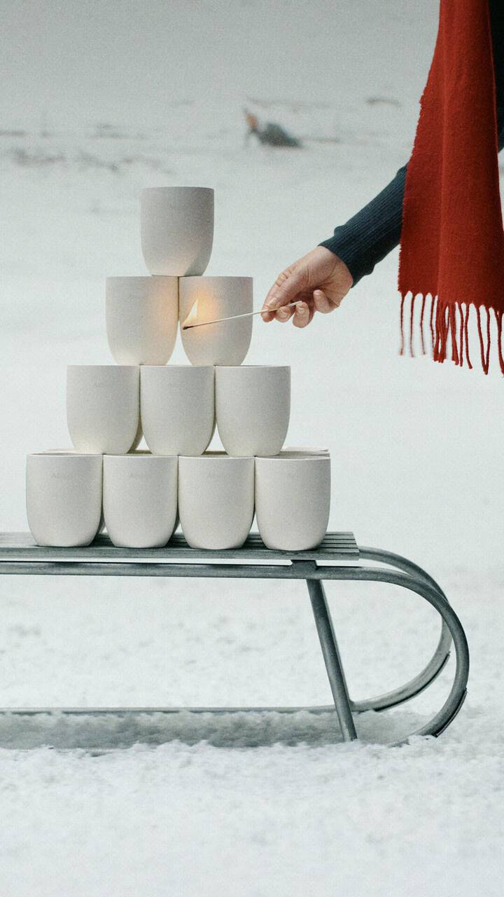 A hand holds a burning match beside white vessels stacked on a metal sledge in snow, with a red fringed scarf to the right.