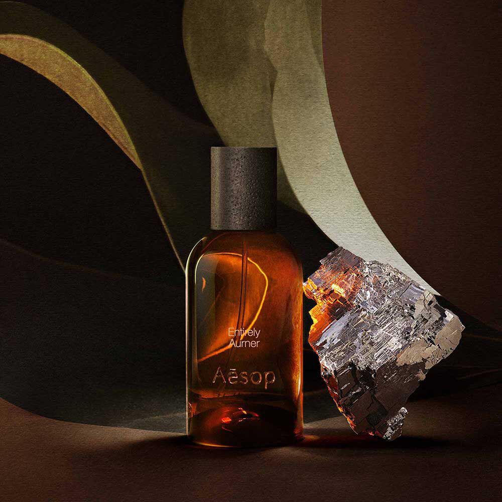 Aesop Entirely Aurner bottle beside a mineral fragment and broad green leaves.
